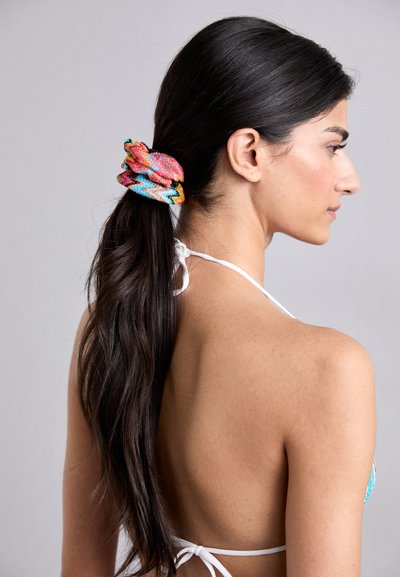 Scrunchie colorat cu un model în zigzag în nuanțe de roz, albastru și galben, realizat dintr-o țesătură texturată. Părul este prins într-o coadă lejeră.