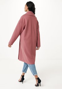 Cappotto rosa oversize e lungo realizzato in tessuto morbido con colletto a scialle, abbinato a jeans azzurri arrotolati e sandali neri con tacco alto.