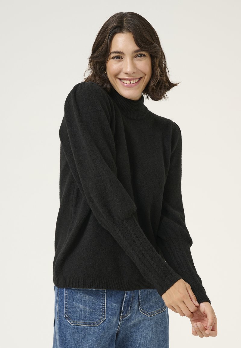 Schwarzer Rollkragenpullover mit gerippten Ärmeln, aus weichem, strukturiertem Material. Kombiniert mit blauen Jeans, präsentiert mit ausgestreckten Armen.