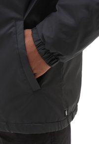 Schwarze gesteppte Jacke mit elastischen Bündchen, einer Seitentasche und einer strukturierten braunen Innenausstattung. Dezentes Logoetikett am Saum.