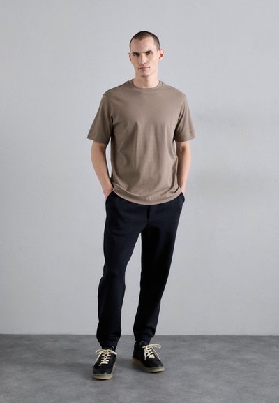 Brun kortermet t-skjorte satt sammen med svarte teksturerte joggebukser og svarte sko med lyse snøring. Enkel, moderne design og avslappet passform.