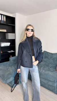 Jachetă bleumarin din lână, cu două rânduri de nasturi, pulover negru cu guler înalt, blugi deschiși la culoare evazați, ținând o geantă neagră. Canapea texturată de culoare turcoaz în fundal.