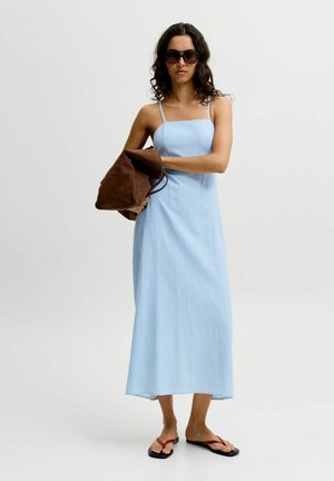 Frau in blauem ärmellosem Kleid und Sonnenbrille hält große braune Tasche, trägt schwarze Flip-Flops vor schlichtem hellem Hintergrund.