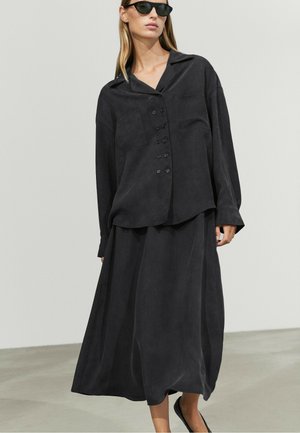 Woman wearing loose black buttoned shirt and long skirt with black sunglasses standing against plain light background.