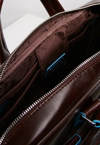 Interno di una borsa in pelle marrone con scomparti con zip, texture liscia, accenti blu e molteplici tasche per l'organizzazione.