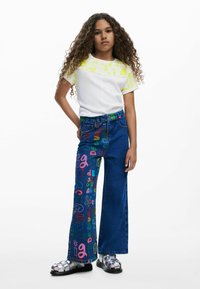 Camisa blanca de manga corta con estampado gráfico amarillo combinada con vaqueros de campana azules que presentan un texto colorido, terminando con sandalias plateadas.