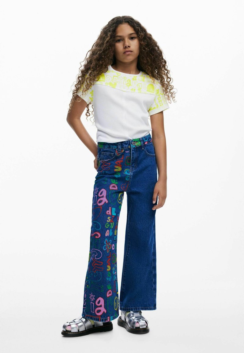 Camisa blanca de manga corta con estampado gráfico amarillo combinada con vaqueros de campana azules que presentan un texto colorido, terminando con sandalias plateadas.