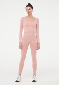 Różowa, prążkowana bluzka z długim rękawem i legginsy z wysokim stanem wykonane z elastycznego materiału, w zestawieniu z białymi butami sportowymi. Płaska faktura, dopasowany krój.