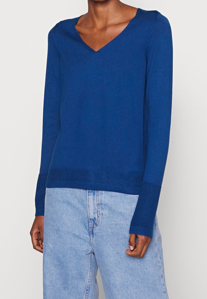 Personne portant un pull bleu foncé à col en V manches longues et un jean en denim bleu clair, debout devant un fond uni.