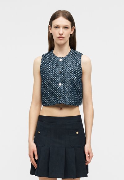 Γυναίκα που φοράει μπλε cropped γιλέκο χωρίς μανίκια με υφή και κουμπιά, καθώς και μαύρη πλισέ φούστα με μπροστινές τσέπες και χρυσά κουμπιά.