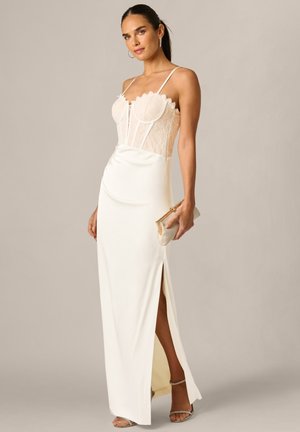 Kobieta w białej sukni z koronkowym gorsetem i rozcięciem na uda, trzyma beżową kopertówkę, pozując na neutralnym tle.