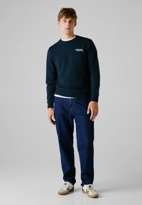 Navy Sweatshirt mit einem weißen Logo, kombiniert mit dunklen blauen Jeans und beigen Sneakers mit schwarzen Akzenten. Glatte Textur, lässiges Design.