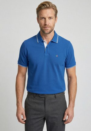 Man met kort blond haar en baard, draagt een blauw poloshirt en donkergrijze broek tegen een effen lichte achtergrond.