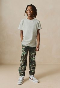 Graues T-Shirt mit kurzen Ärmeln kombiniert mit grünen Camo-Cargohosen mit elastischen Bündchen und Seitentaschen. Abgerundet wird das Outfit mit weißen Turnschuhen.