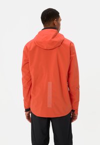 Man die met zijn rug naar de kijker staat, gekleed in een felle oranje hoodie met reflecterende stippen op de onderrug en zwarte broek.