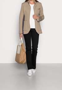 Mujer con blazer beige sobre camisa blanca, pantalones negros, zapatillas blancas, llevando un gran bolso de mano beige y marrón, de pie sobre un suelo gris.
