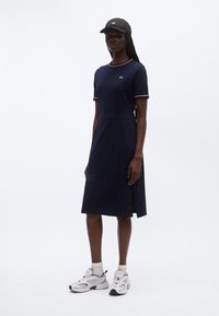 Tumši zila kokvilnas kleita ar īsām piedurknēm, rozā apdare kaklam un manžetēm, sānu šķēlums, kopā ar melnu cepuri un baltām sporta kurpēm.