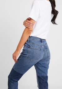 Jeans de denim de tiro alto con un diseño de patchwork que incluye secciones de azul claro y oscuro. Cinco bolsillos y un cierre estándar con cremallera.