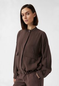 Braune, leichte Jacke mit rundem Ausschnitt, elastischem Saum und zwei Vordertaschen. Verfügt über eine subtile Textur und ein lässiges Design.