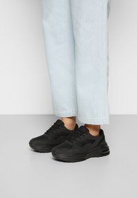 Chaussures de sport noires avec panneaux en mesh, bout arrondi et semelle épaisse avec amorti à air, portées avec un jean bleu clair raccourci.
