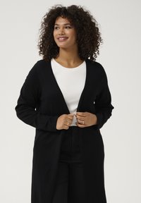 Cardigan nero a maglia con maniche lunghe, apertura frontale e una vestibilità rilassata, indossato sopra una maglietta bianca. Texture liscia e dettagli minimi.