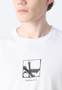 Camiseta de algodón blanca con un diseño gráfico negro y el texto "Calvin Klein Jeans" en la parte inferior, con cuello redondo y mangas cortas.