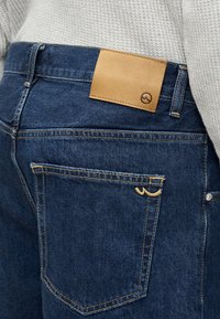 Τσέπη πίσω από τζιν από σκούρο μπλε denim με κίτρινη ραφή και ταμπέλα από δέρμα σε ταμπά χρώμα, φορεμένα με ανοιχτό γκρι πουλόβερ με υφή.