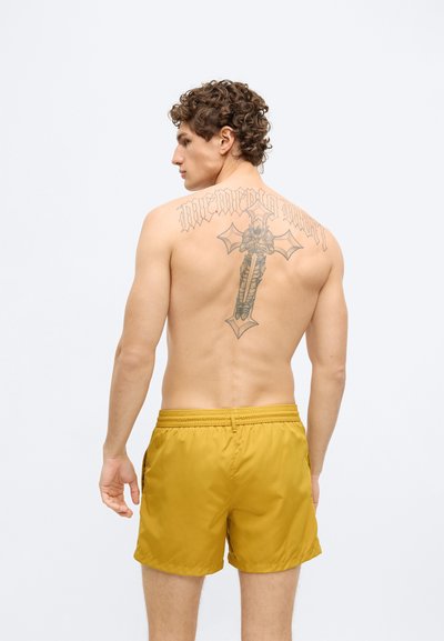 Sárga fürdőnadrág elasztikus derékpánttal, könnyű anyagból. A hátsó részén nagy fekete tetoválásminta található.