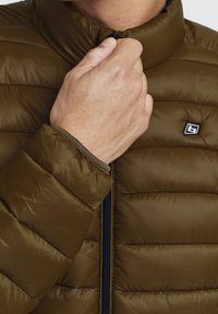 Olivengrøn puffjakke med høj krave, der viser en quiltet tekstur. Har lynlås og et mærket logo på øverste venstre side.