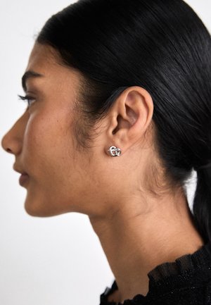 Profil d'une femme aux cheveux foncés attachés, portant une boucle d'oreille clou en forme de cœur argentée et un haut noir avec un col à volants.