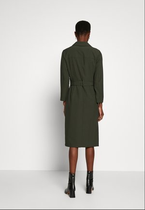 Person iført en knælængde, olivengrøn frakke med bælte og lange ærmer, stående med ryggen til, kombineret med sorte ankelstøvler med hæle.