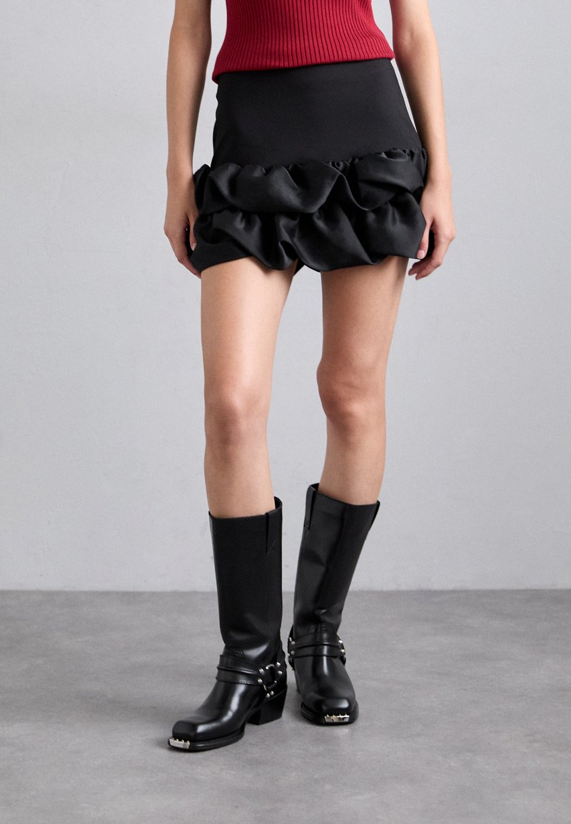 Mini-jupe noire avec un ourlet en satin froncé, associée à des bottes en cuir noir montantes avec des détails en métal argenté et un talon bloc.