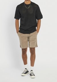Homme portant un pull à manches courtes noir texturé, un short beige, des chaussettes blanches, des baskets noires et blanches, et un collier fin en or, se tenant les mains dans les poches.