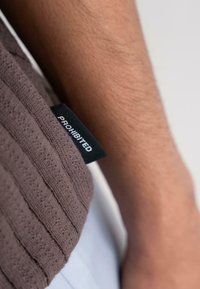Brown patterned fabric with vertical stripes and a black tag that reads "PROHIBITED," partially visible against a skin-toned arm.