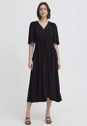 Donna con un abito midi nero a maniche corte, con bottoni sul davanti, cintura in vita e sandali neri con tacco e punta aperta, in piedi su uno sfondo semplice.