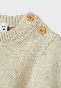 Maglione lavorato a maglia beige chiaro con colletto a coste e due bottoni in legno sulla spalla. Texture morbida e design casual. Etichetta della taglia visibile.