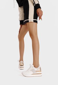 Sneakers atletici bianchi con accenti dorati e dettagli in rete, caratterizzati da una suola chunky e pannelli in tessuto texturizzato.