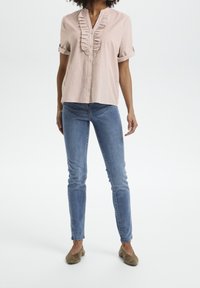 Camicia rosa chiaro a maniche corte con dettagli arricciati sul davanti, abbinata a jeans blu e ballerine marroni. Tessuto liscio, vestibilità rilassata.