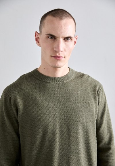 Pull en maille vert olive avec un motif texturé, doté d'un col rond et de manches longues, sur un fond neutre.