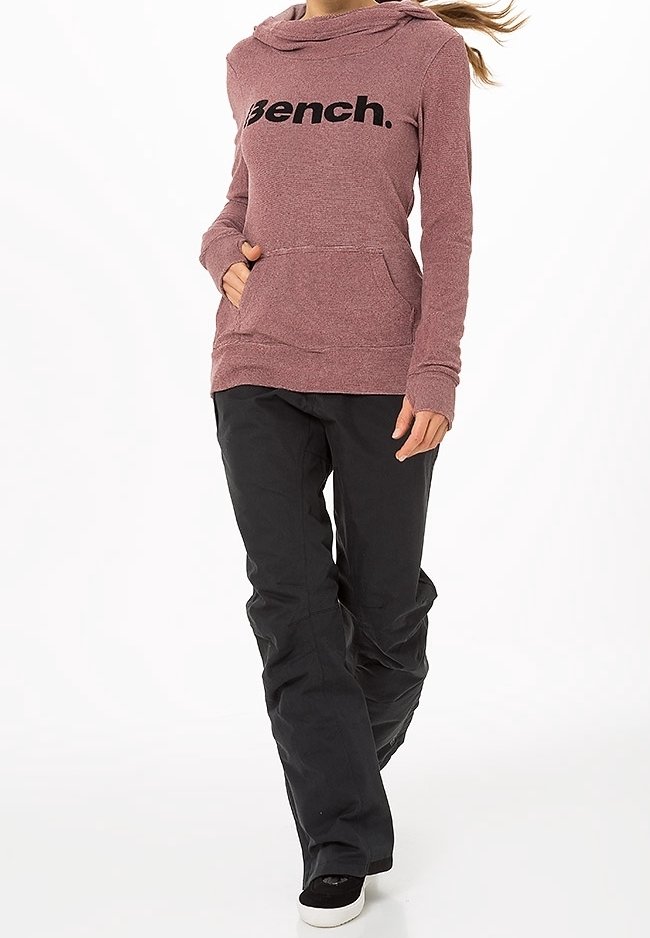 Femme portant un hoodie Bench rose poussiéreux avec poche avant et pantalon noir ample, marchant vers l'avant sur un fond blanc.