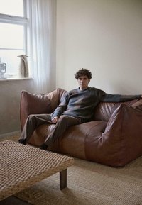 Braunes Leder-Bohnensofa mit weicher Textur; grauer gemusterter Pullover, getragen von einer sitzenden Person, begleitet von einem geflochtenen Couchtisch.