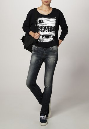 Person trägt einen schwarzen Sweatshirt mit dem Aufdruck „In Skate We Trust“, verblasste dunkle Jeans, marineblaue Sneakers und trägt einen schwarzen Rucksack auf einer Schulter.