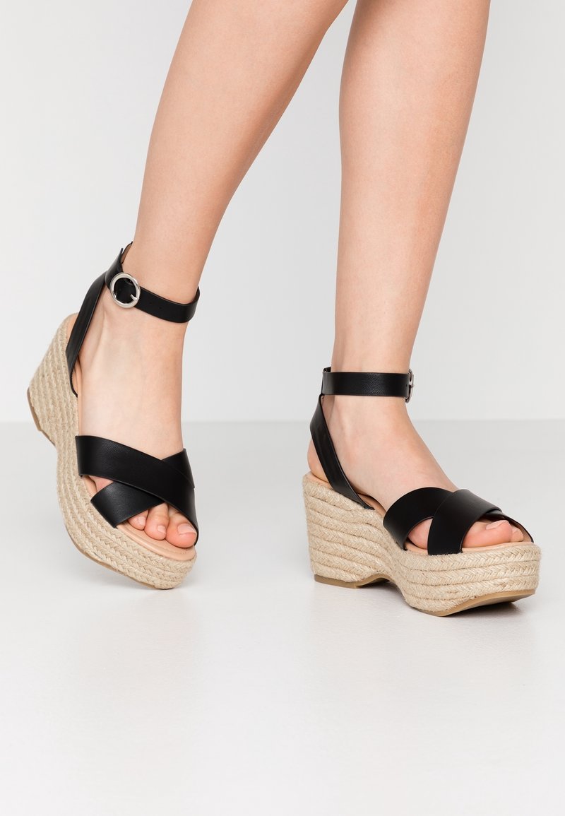 Pieds portant des sandales compensées en cuir noir avec bride cheville et talons plateforme en corde tressée sur fond clair.