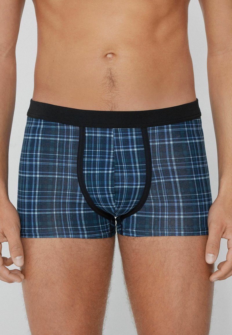 Mužský trup a horní stehna oblečené v modrých a černých kostičkovaných boxerkách s černým pasem a lemem.