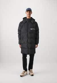 Svart dunjakke med glidelås foran, hette og sidelommer. Har logo på brystet, teksturerte paneler og oversized passform. Kombinert med mørke jeans og sko.