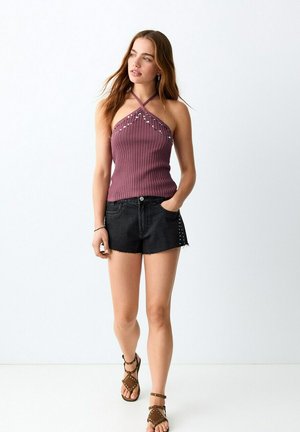 Jeune femme marchant, portant un haut dos nu violet avec des clous, un short en jean noir et des sandales marron cloutées sur un fond blanc uni.