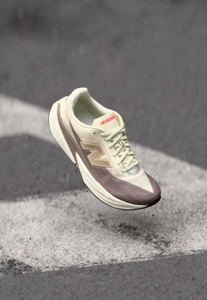 Lyse beige og brune New Balance løbesko med snørebånd, svævende i luften over asfalt med hvide vejmarkeringer.