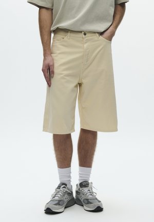 Pantaloncini beige fino al ginocchio indossati con calzini bianchi e scarpe da ginnastica grigie su una persona in piedi con una mano in tasca.