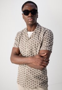 Camisa de manga curta, beige, com um padrão de design circular, apresentando um fecho de botão. Modelo a usar óculos de sol pretos, com os braços cruzados.