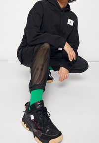 Zwarte hoodie met een logo, zwarte mesh broek en stevige zwarte sneakers met groene en rode accenten. Groene sokken zichtbaar boven de enkel.